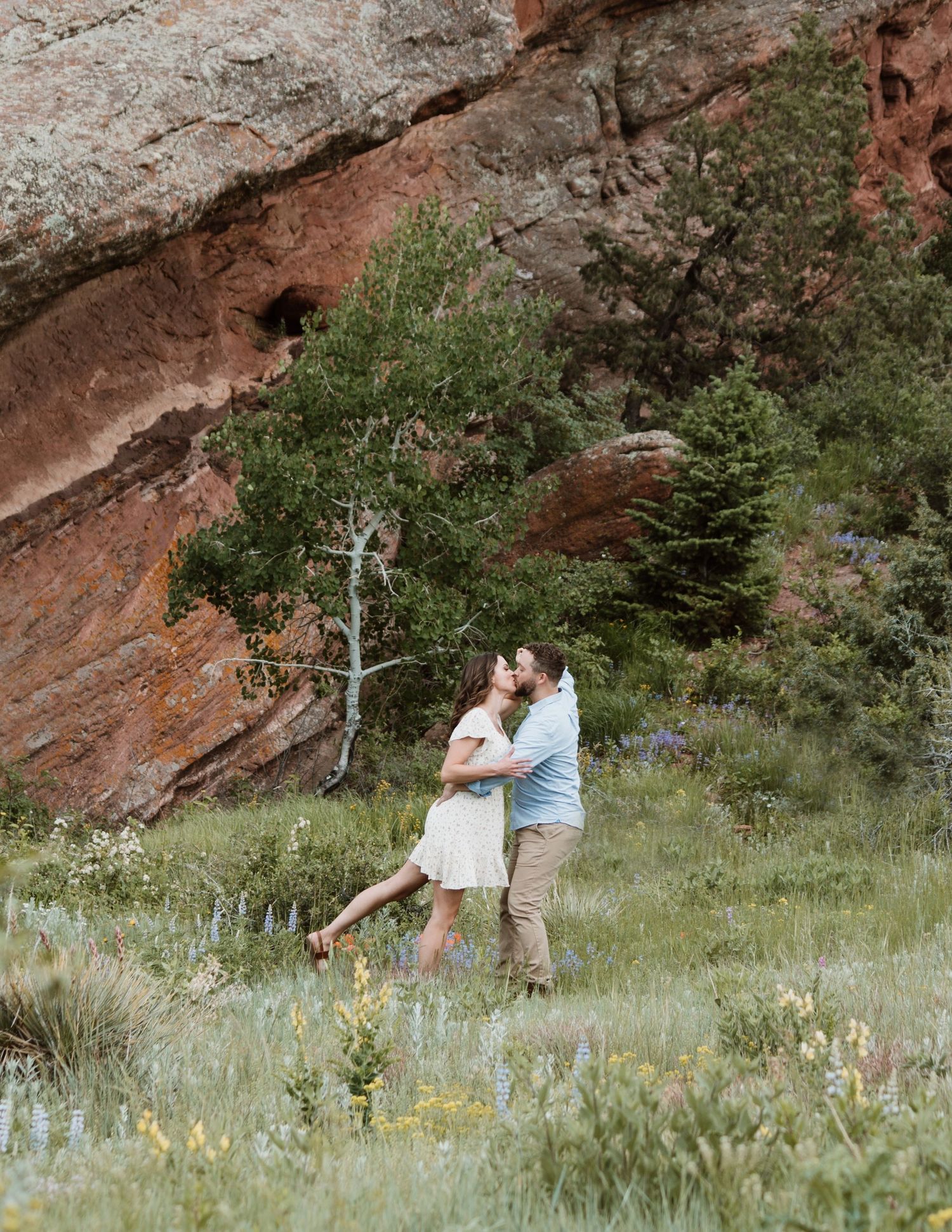 A romantic couple embraces in front of dramatic red rock formations and greenery.