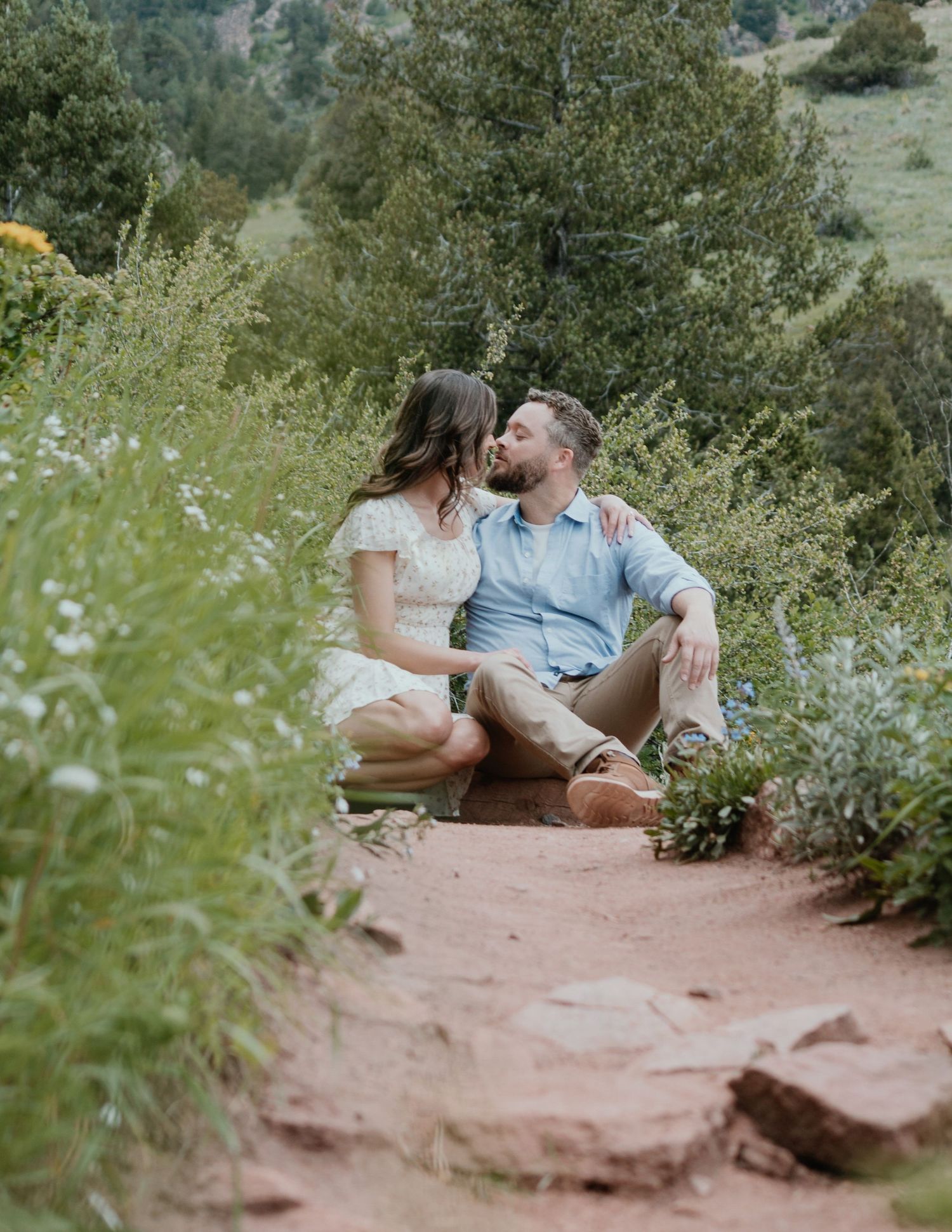 A couple sits together on a red rock trail surrounded by wildflowers and mountain scenery.