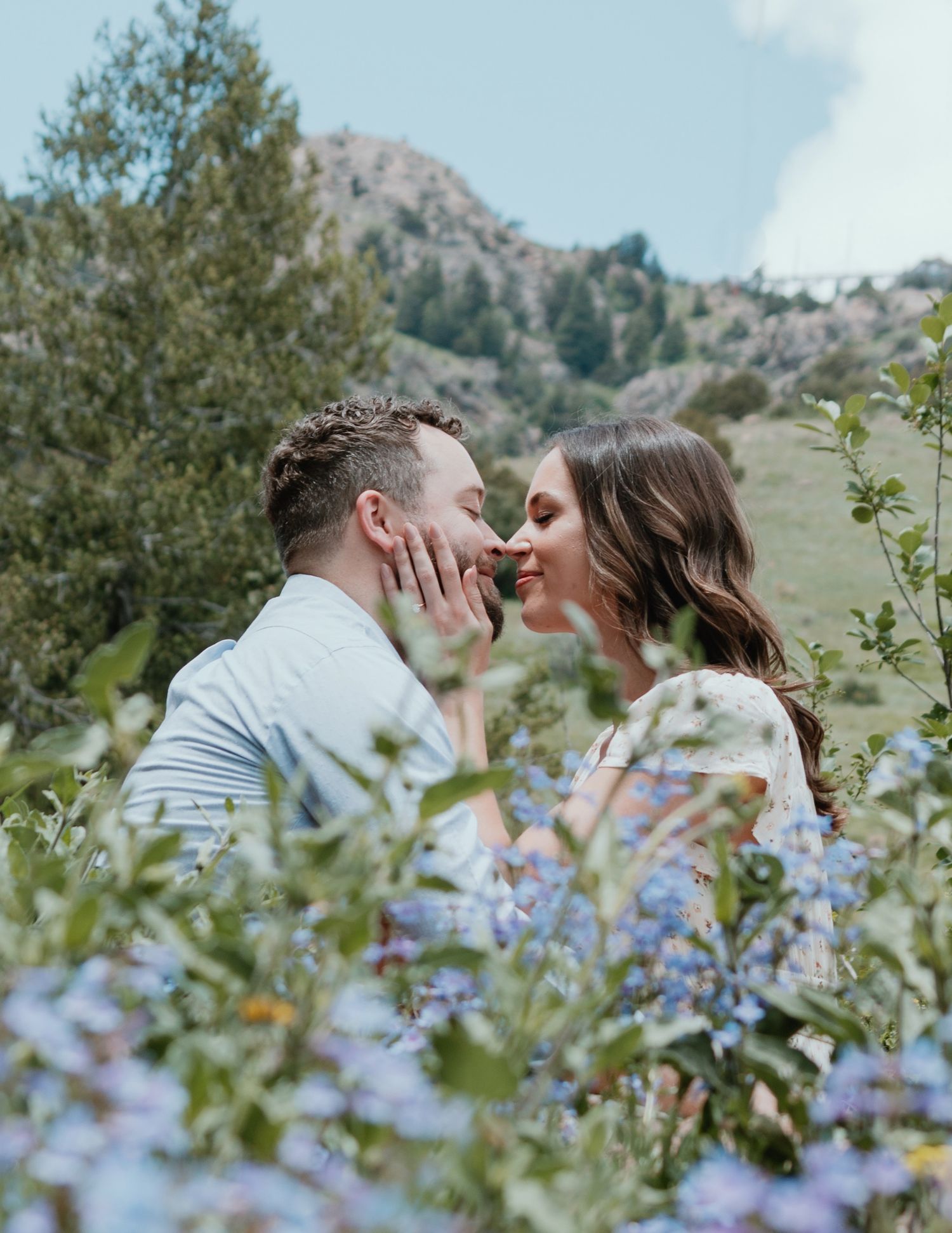 A tender moment between a couple surrounded by blue wildflowers and mountain scenery.