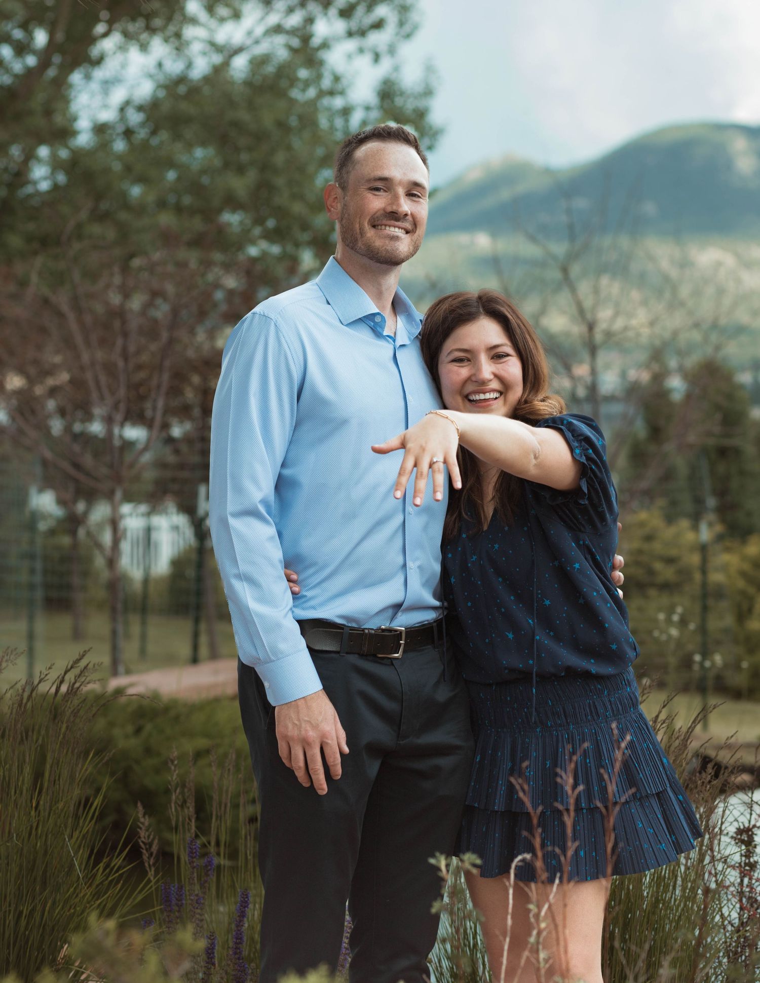 A happy couple showing off an engagement ring with mountains in the background.