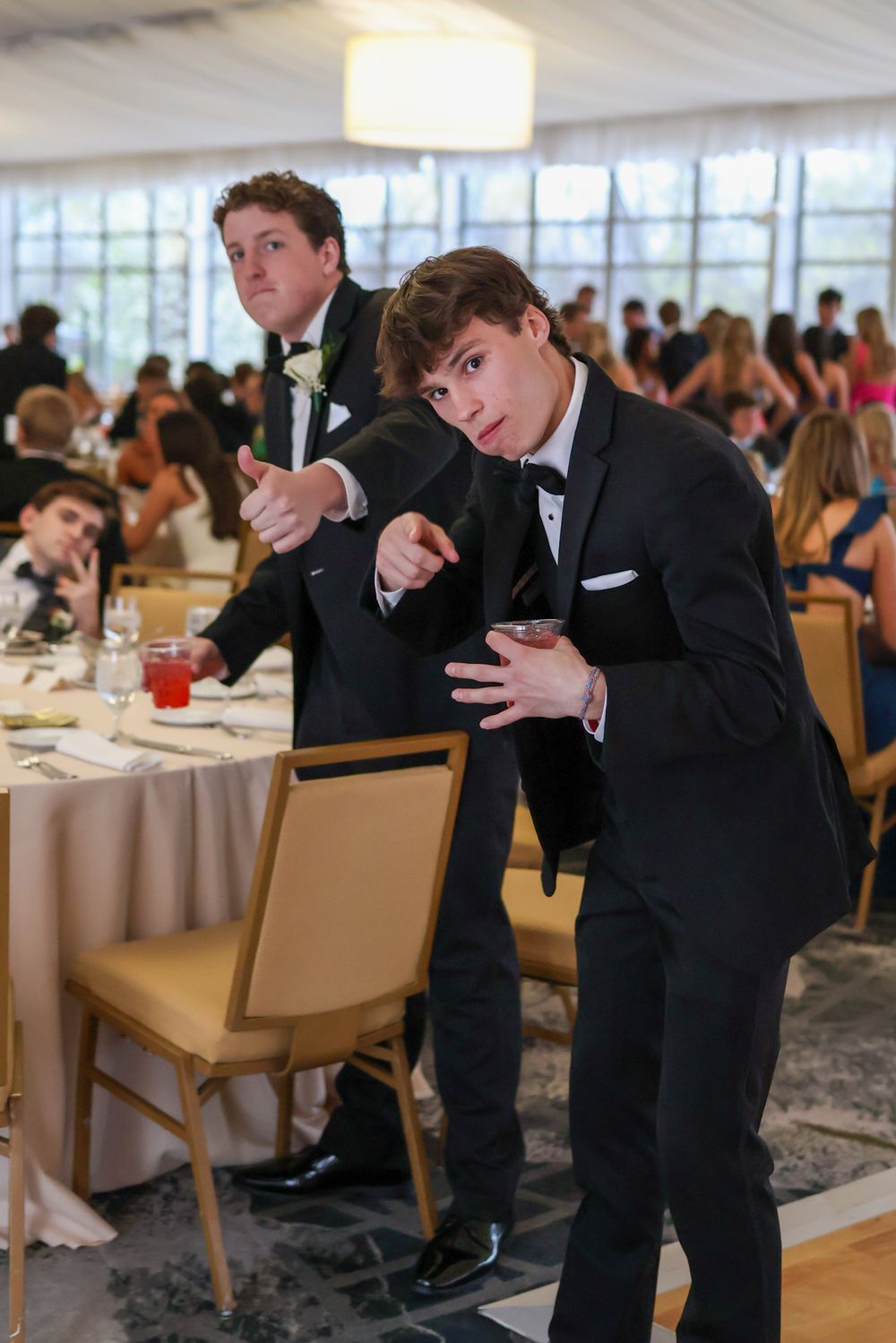 Young men in formal attire strike playful poses at their seats during a banquet event.