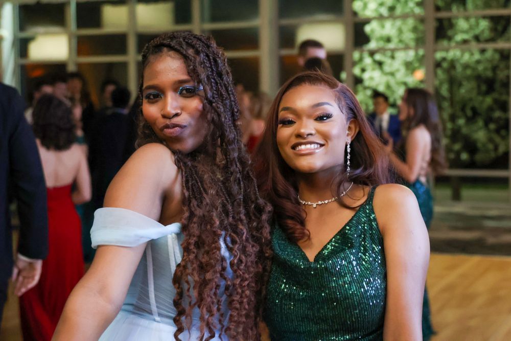 Two formal attendees pose together at a special event, one in a white dress and one in a sparkling green gown.