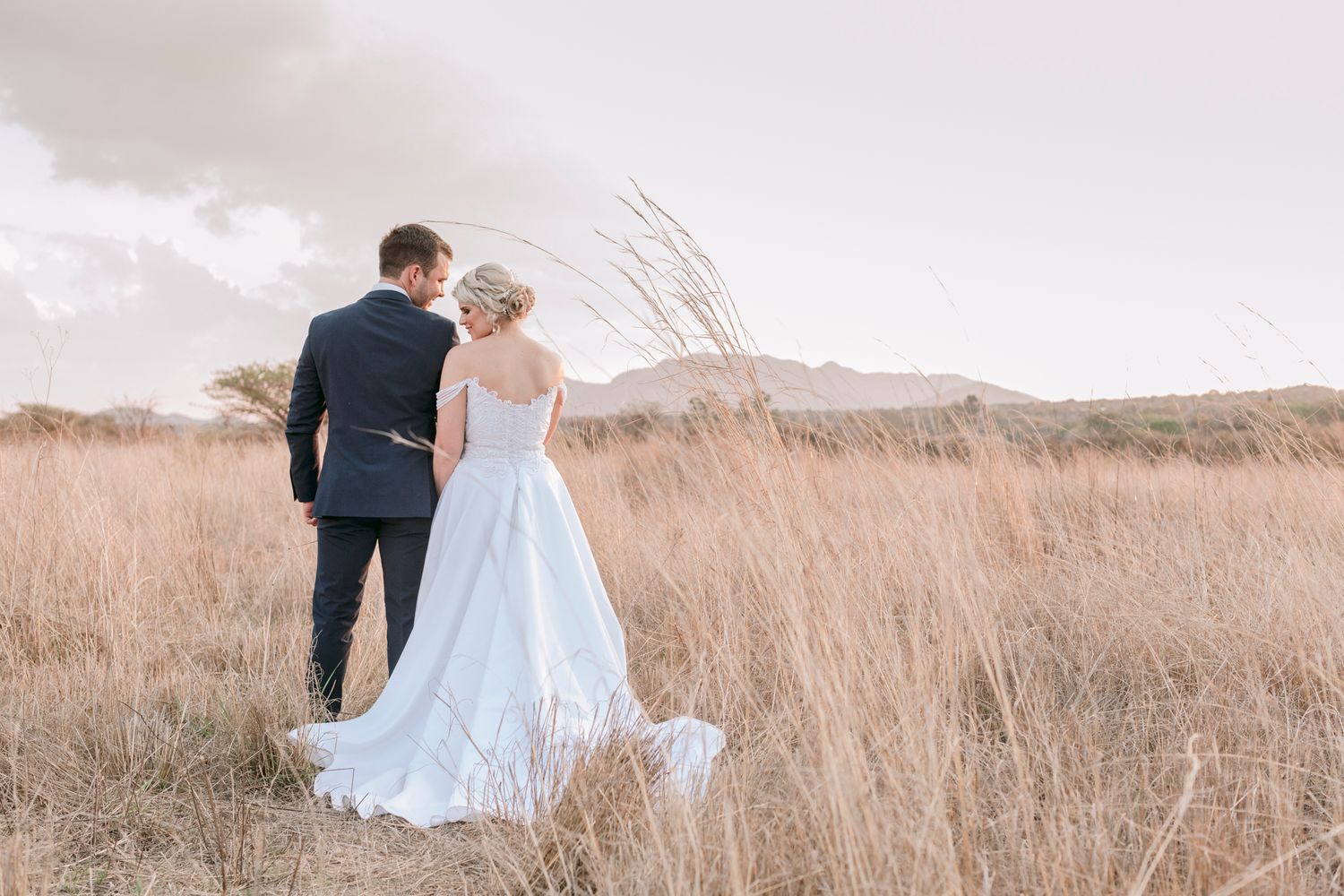 Bride and groom walking through golden grass field with mountains behind.