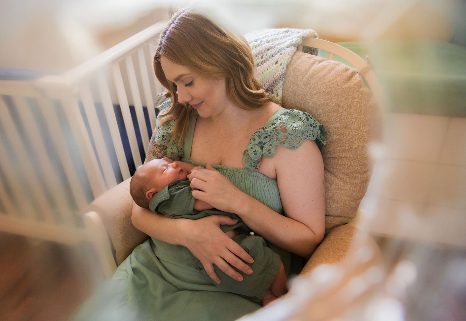 A woman in a sage green dress sits in a rocking chair during a tender nursing session with her baby.