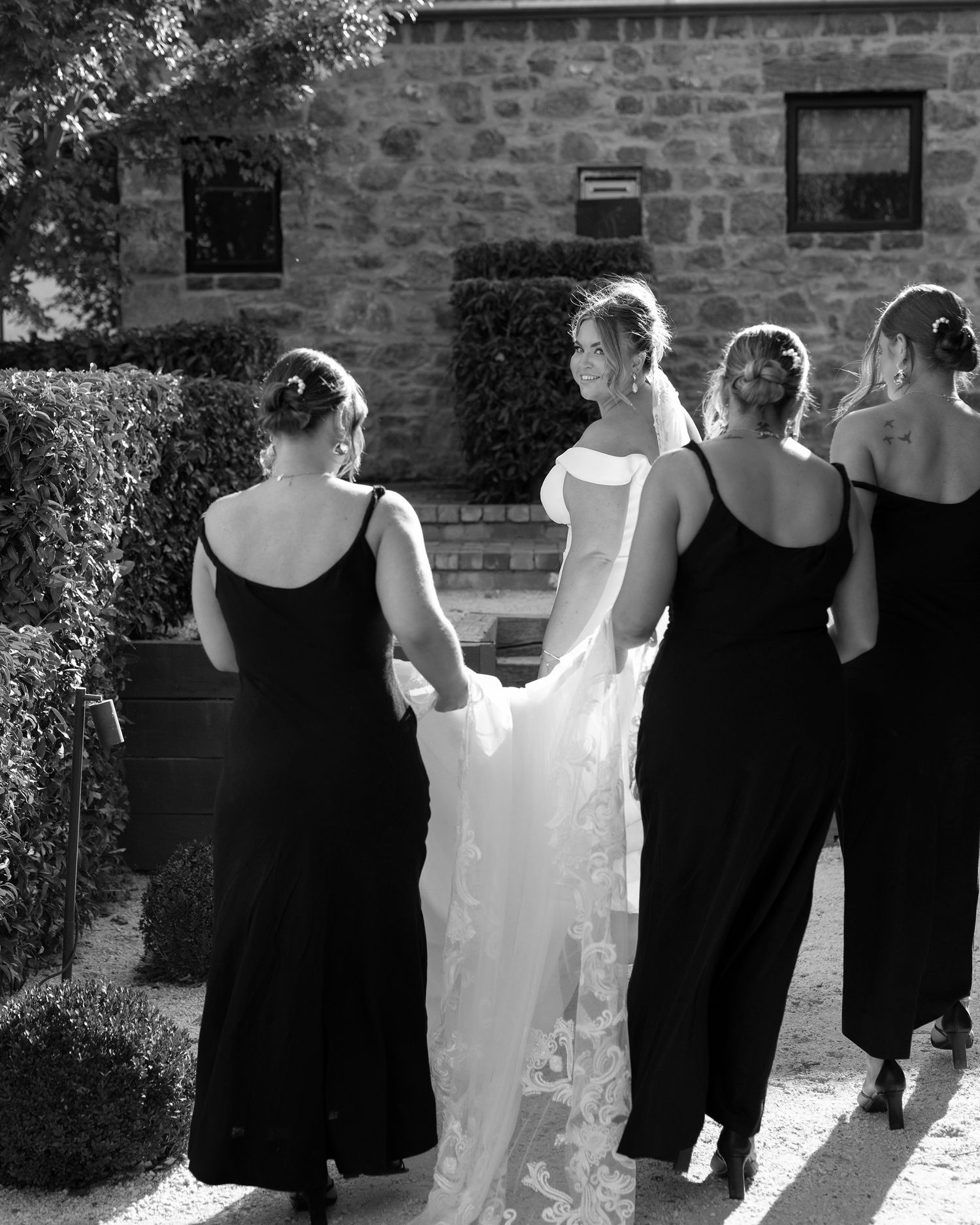 Black and white photo of a bride in a lace gown with bridesmaids in black dresses walking toward a stone building.