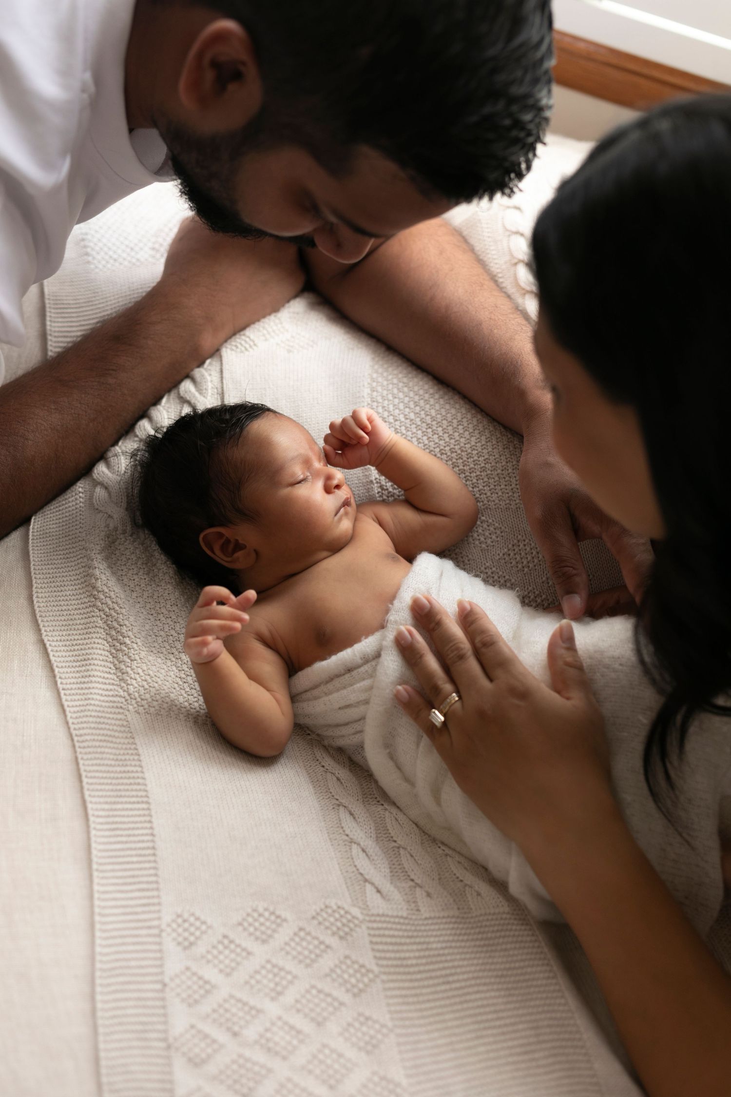 Parents lovingly gaze at their sleeping newborn baby wrapped in a white knit blanket.
