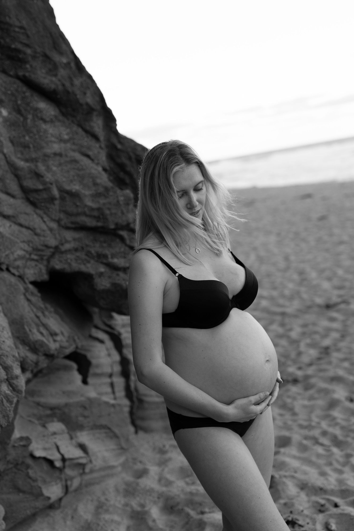 Pregnant woman in black bikini cradling her baby bump on a sandy beach near rocky cliffs in black and white photo.