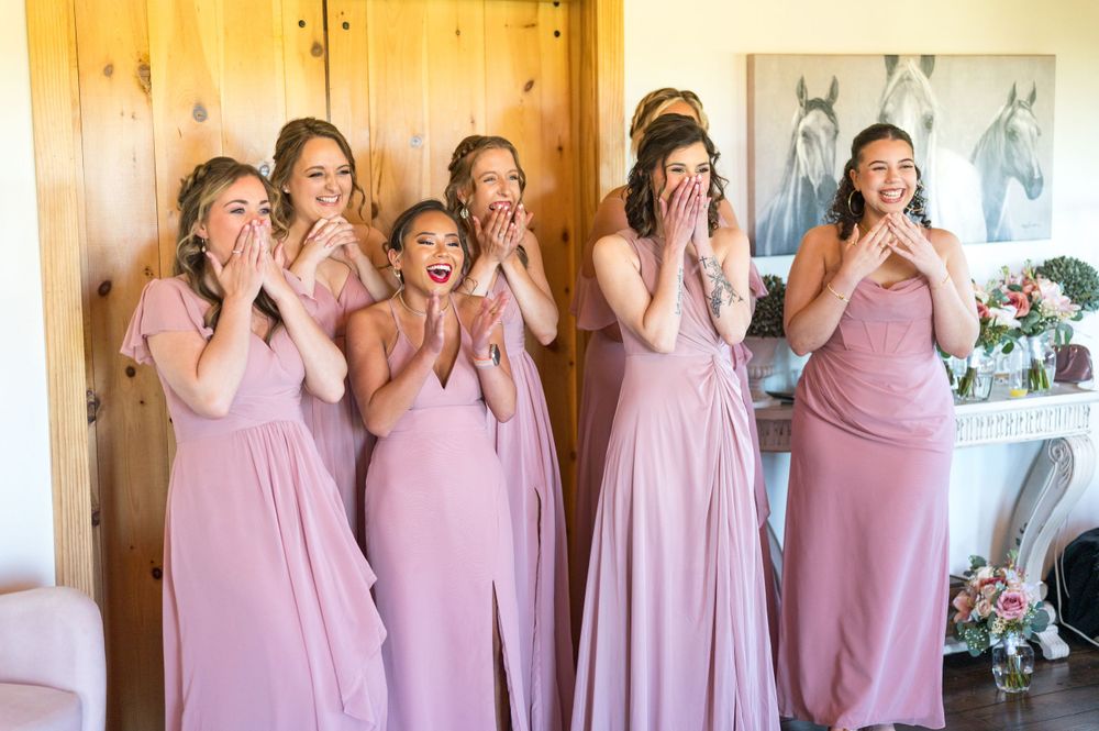 Bridesmaids in dusty rose dresses react with surprised expressions during a wedding day moment.