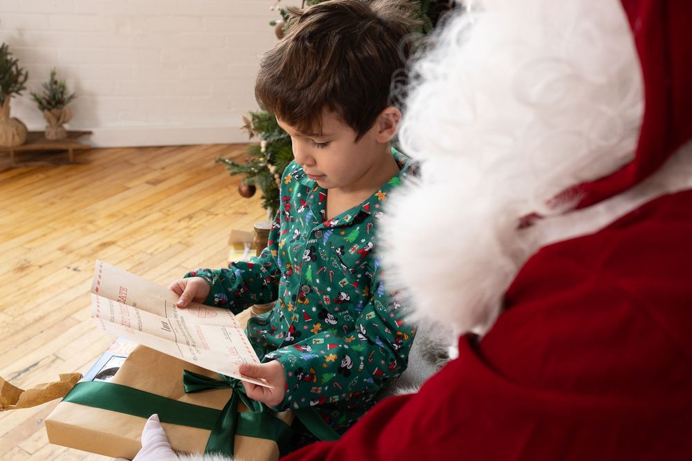 A young child in a green holiday sweater writes a letter to Santa while sitting at a wooden table.