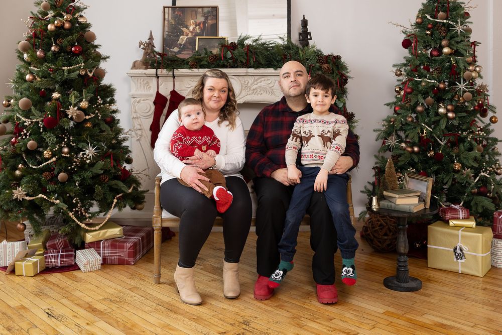 A family sits between two decorated Christmas trees with wrapped presents on a hardwood floor during a holiday photo session.