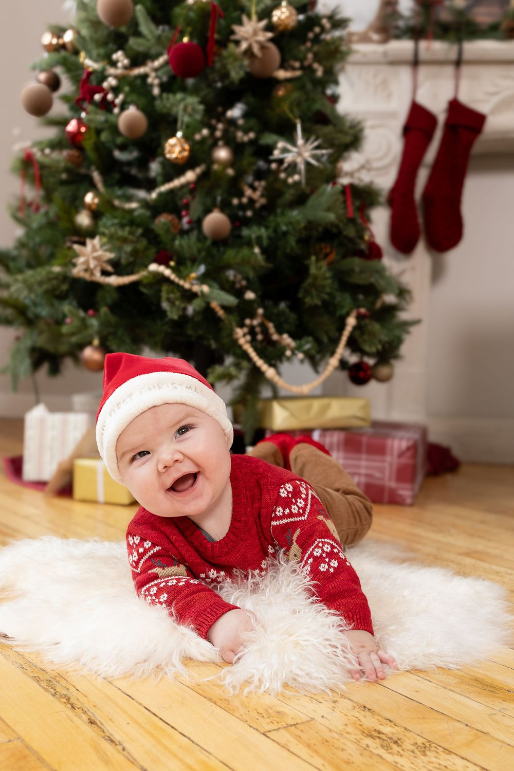 Baby in red Christmas outfit and Santa hat laying on white fur rug near decorated Christmas tree with presents.