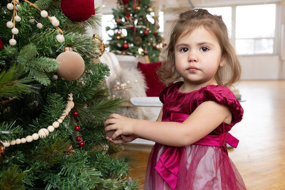 A young child in a burgundy velvet dress helps decorate a Christmas tree with ornaments and garland.