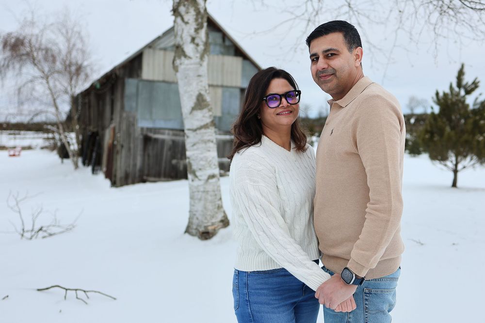 A couple stands close together in a snowy winter landscape with rustic barns and buildings in the background.