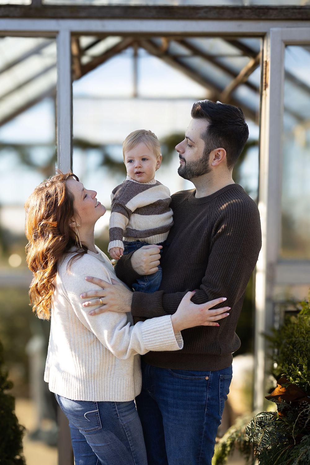 Family shares tender moments together in a bright greenhouse setting with natural sunlight streaming through glass walls.