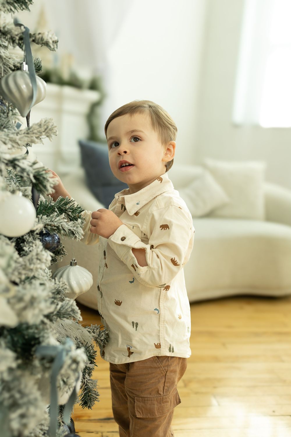 A toddler in a beige sweater and brown pants stands next to a white Christmas tree in a bright living room.