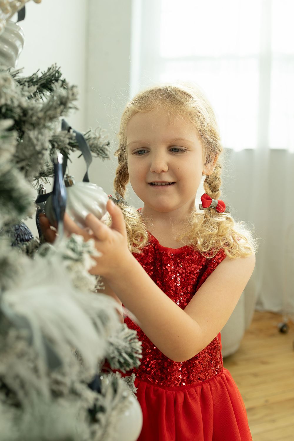 A small child in a sparkly red holiday dress decorates a frosted Christmas tree with ornaments.