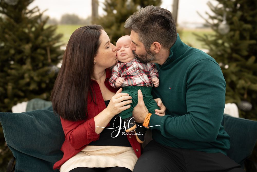 A couple in red and green sweaters embraces outdoors among evergreen trees in a romantic winter setting.