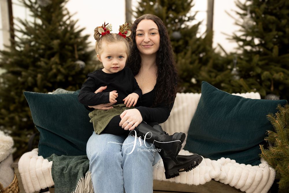 A mother and child sit together on a couch outdoors surrounded by evergreen trees in a winter setting.