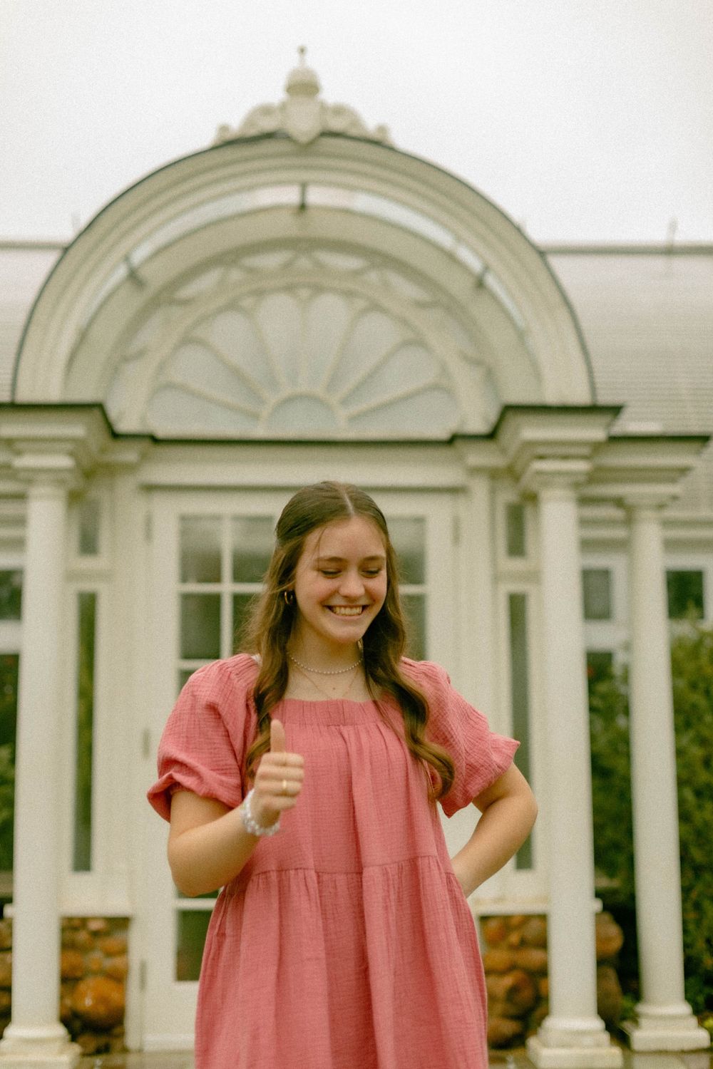 Graduate in pink dress poses joyfully in front of white architectural columns.