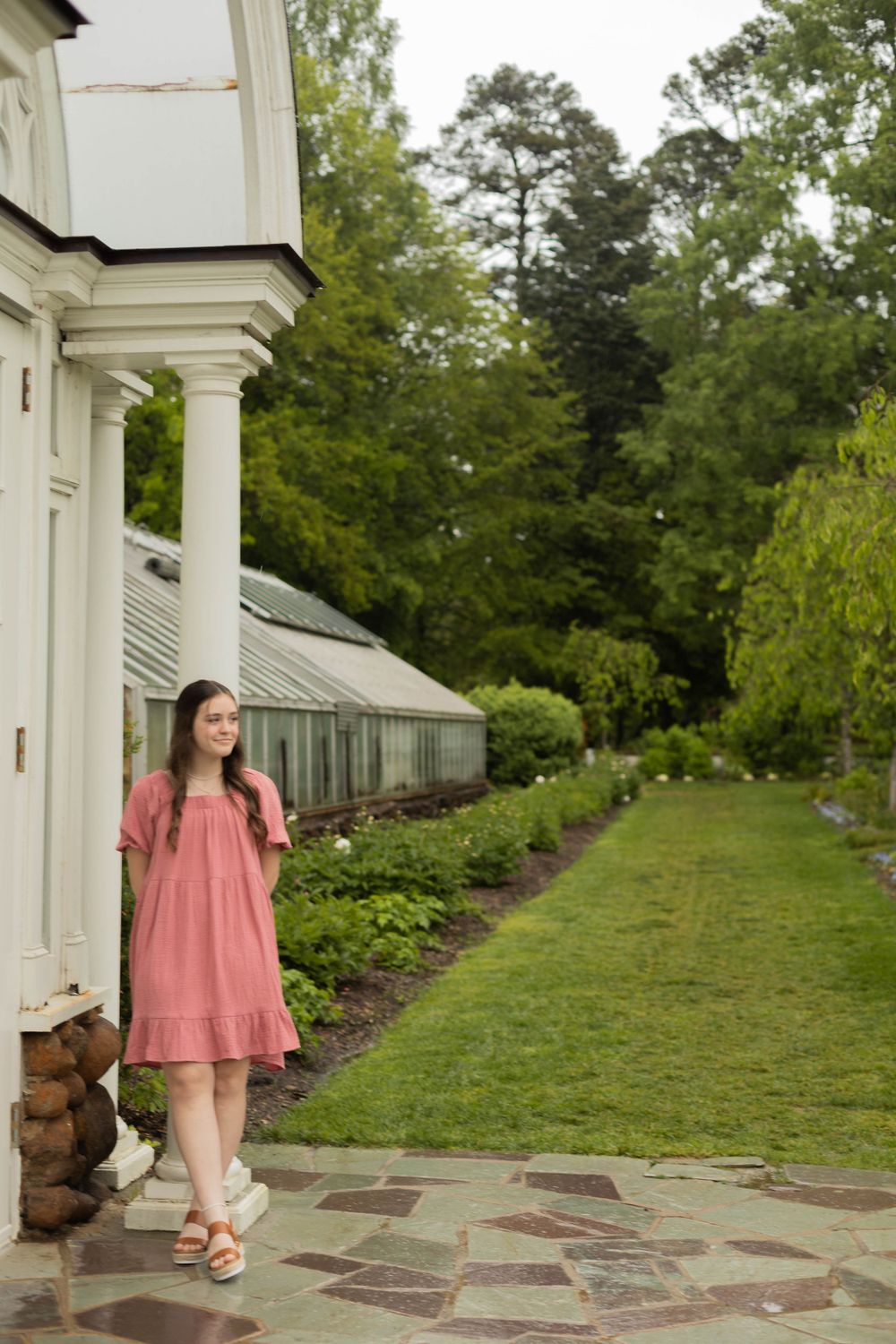 Pink dress stands out against white columns and lush garden landscape with greenhouse in background.