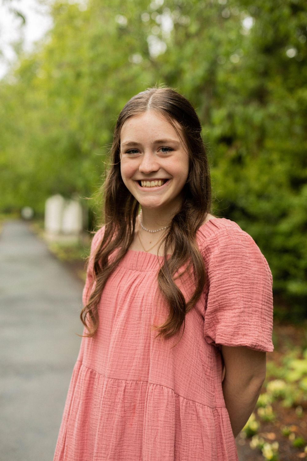 Natural portrait on garden path with soft pink dress against lush green background.