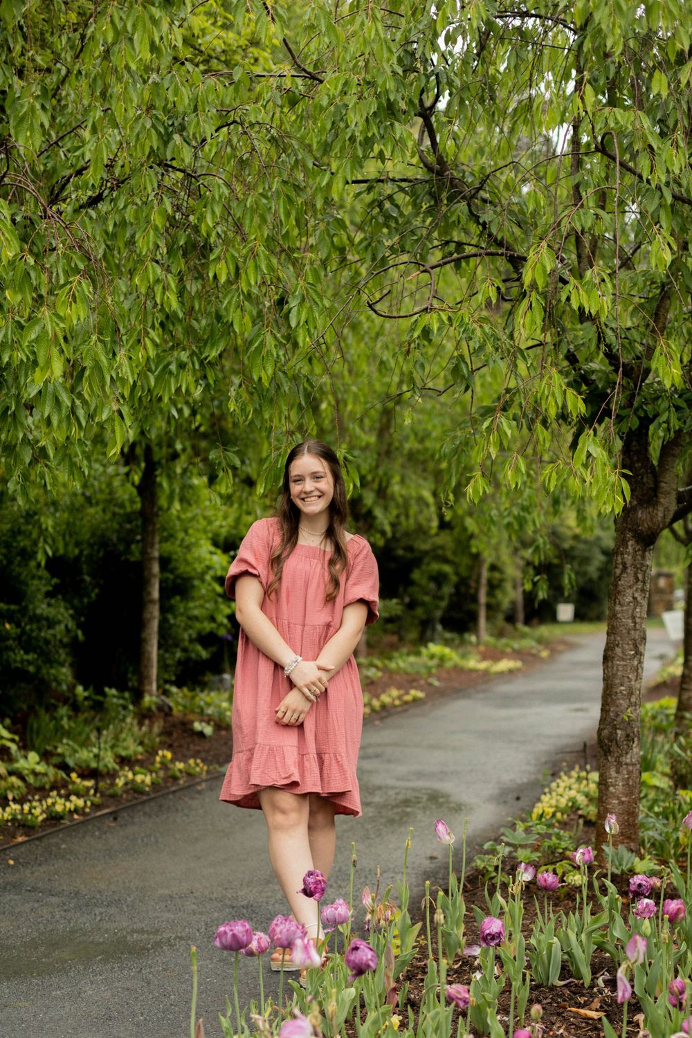A person in a coral dress stands on a garden path surrounded by lush greenery and pink flowers.