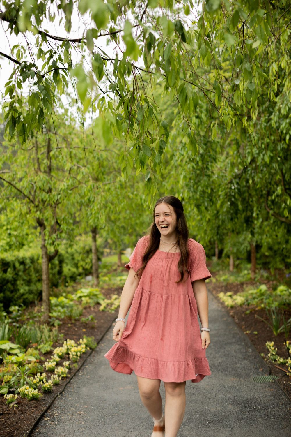 A person in a pink dress laughs joyfully while walking along a garden path under willow trees.