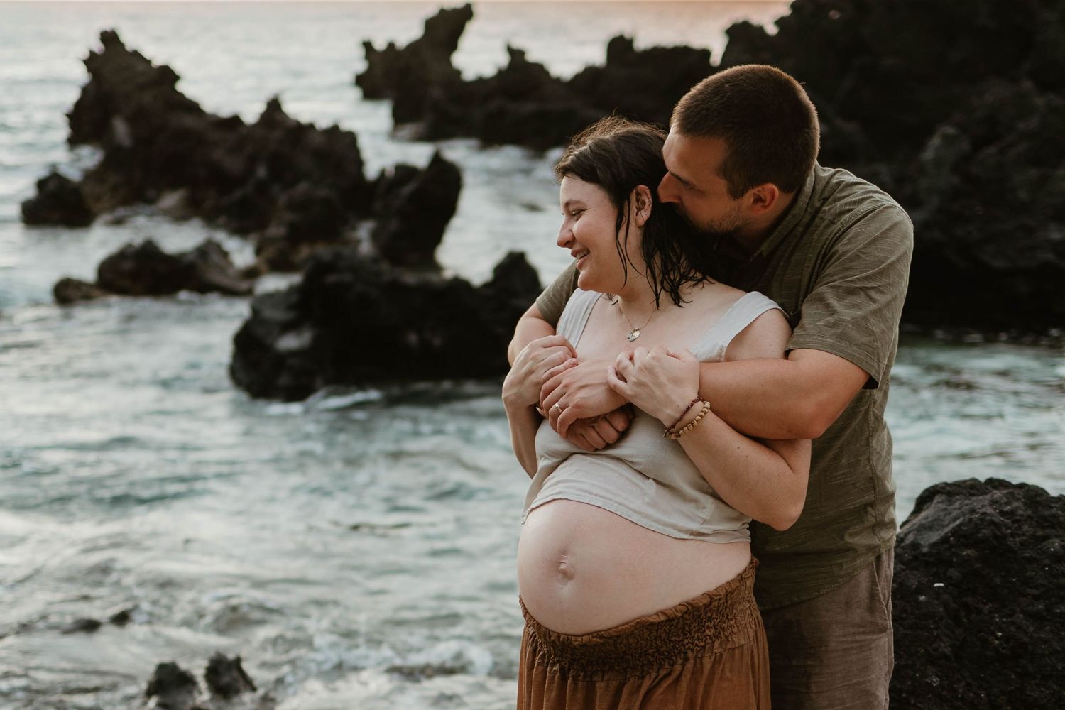 Expectant couple embracing on rocky beach during sunset with waves crashing behind them.