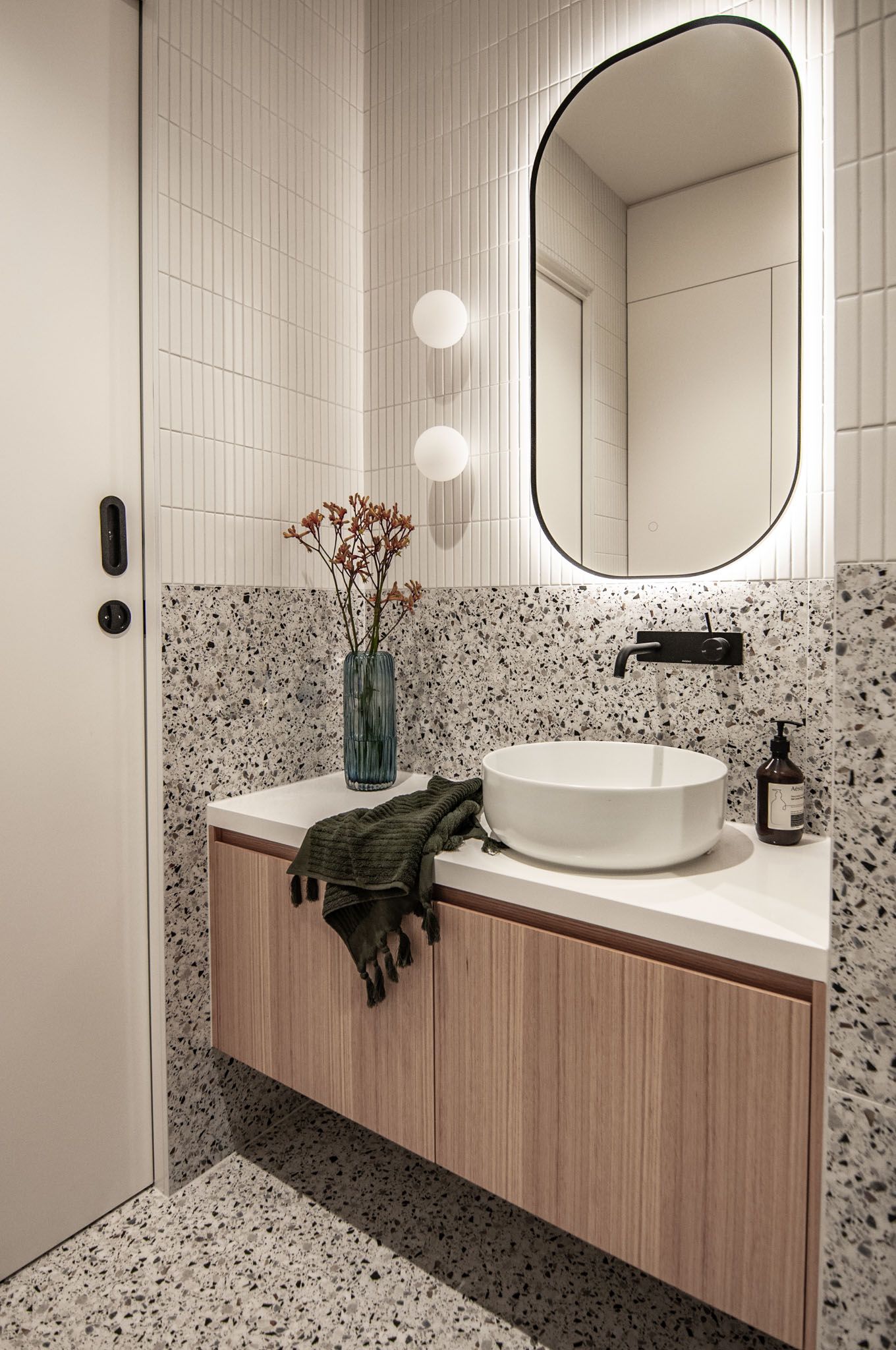 Modern bathroom featuring terrazzo tiles, oval mirror, floating vanity, and sleek lighting fixtures.