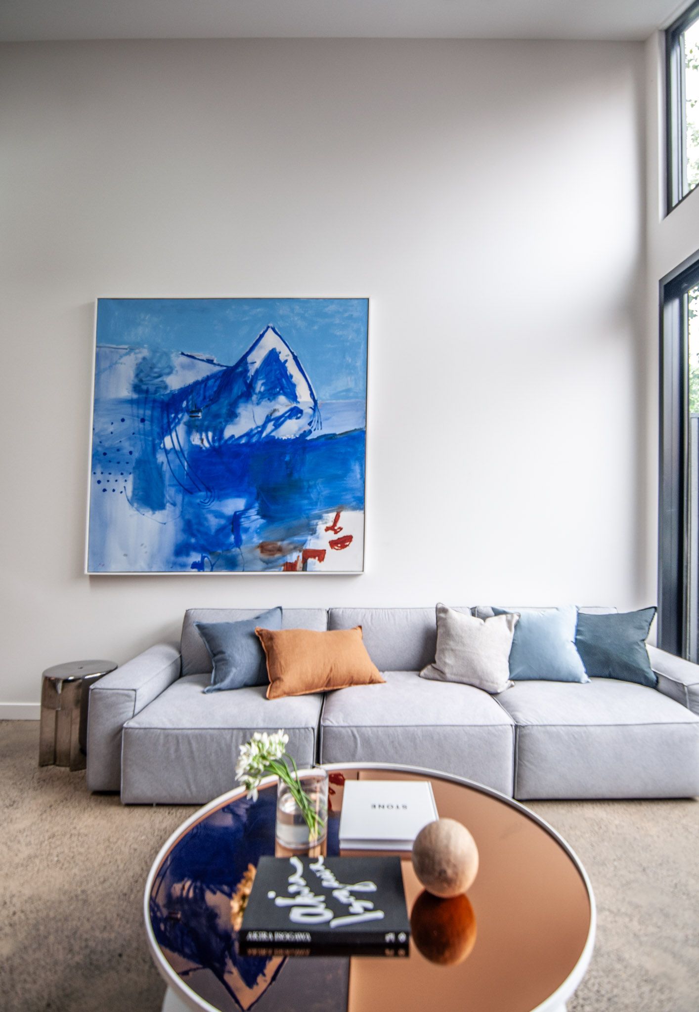 Contemporary living room with low-profile gray sofa, blue abstract art, and round copper coffee table.