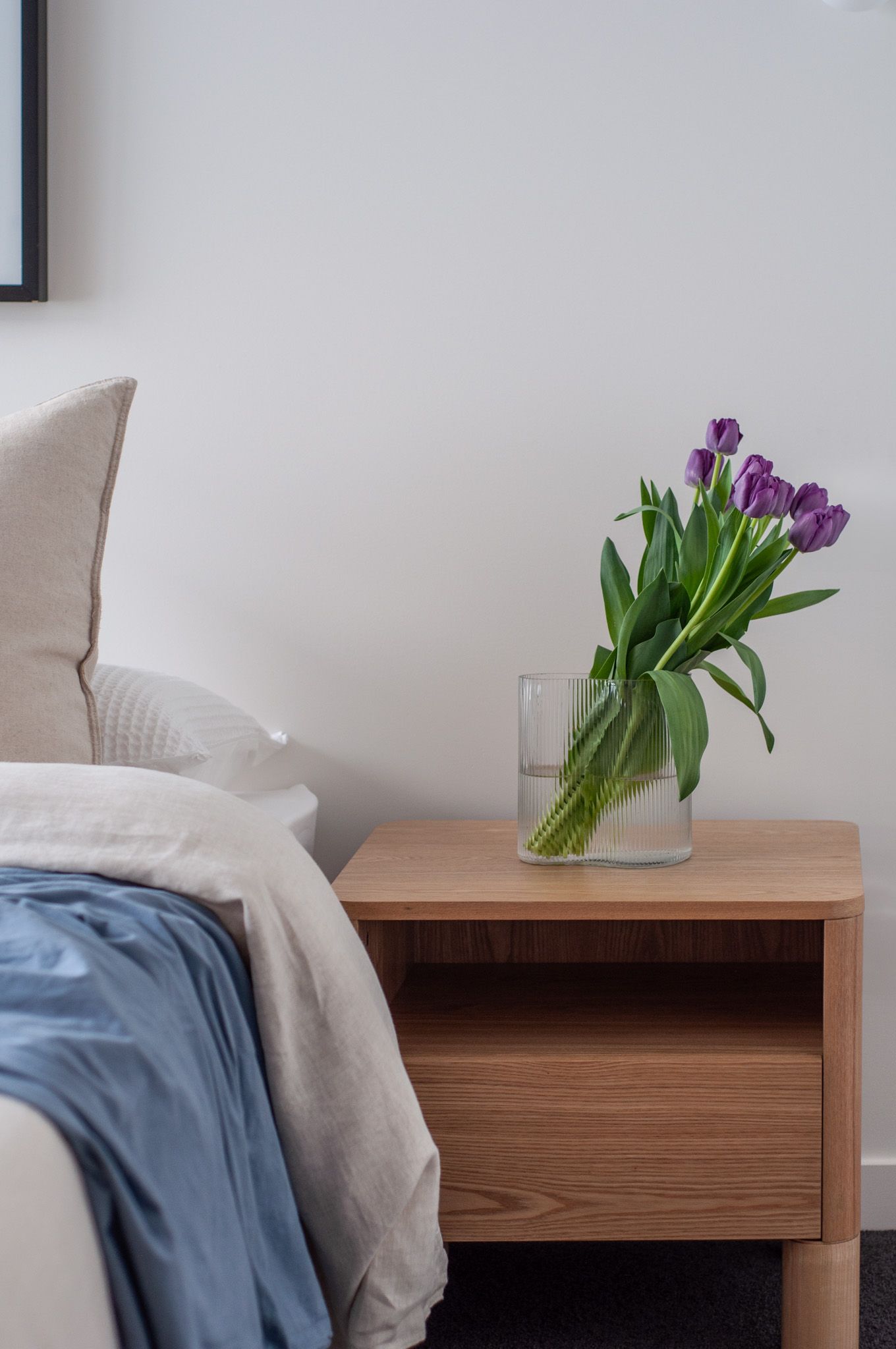 Minimalist bedside table with purple tulips in glass vase against neutral bedding.