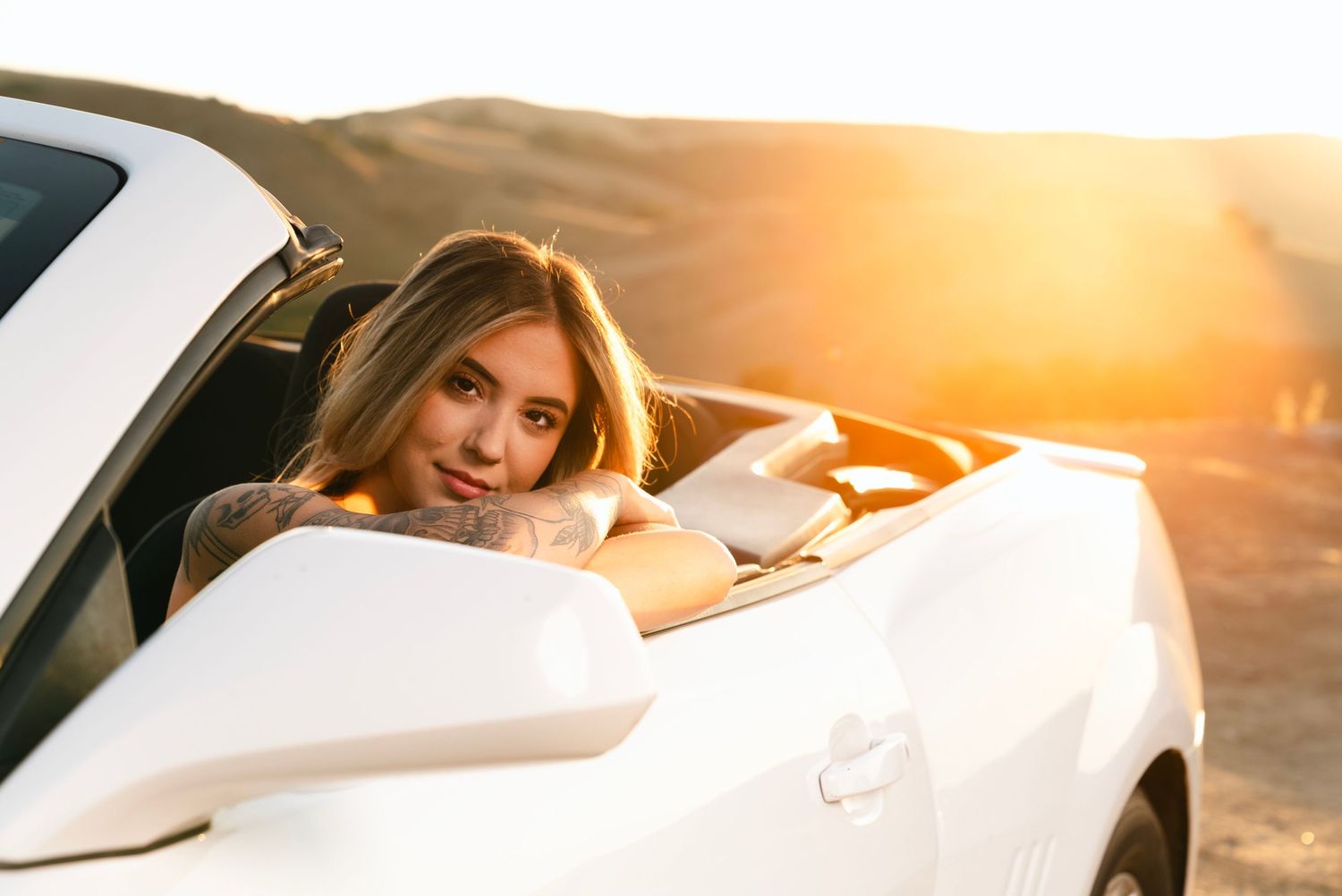 a young woman in a red top and blue jeans poses inside of her white camaro on a hill at sunset