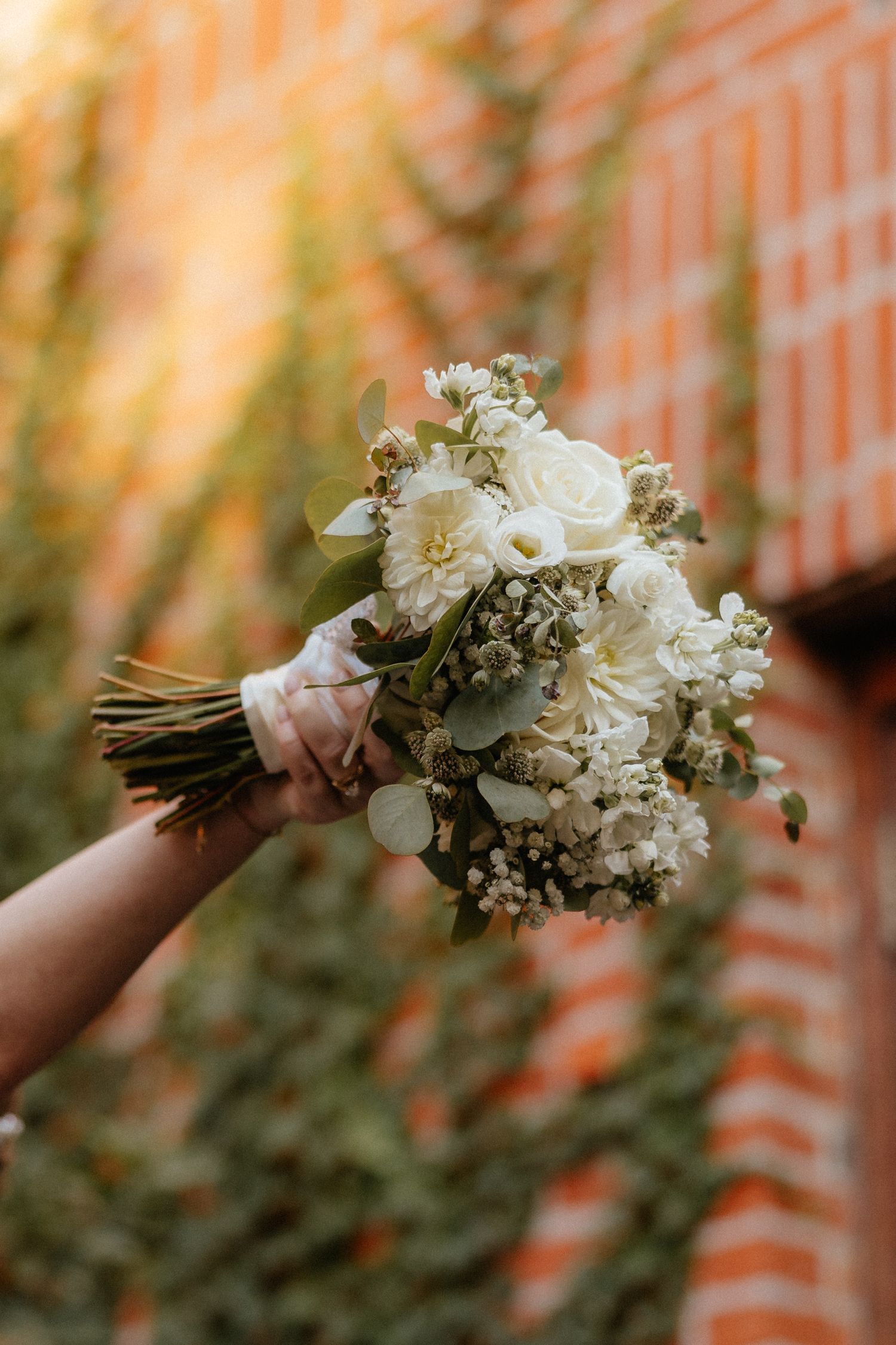 A delicate white and cream bridal bouquet with roses and baby's breath against a brick backdrop.