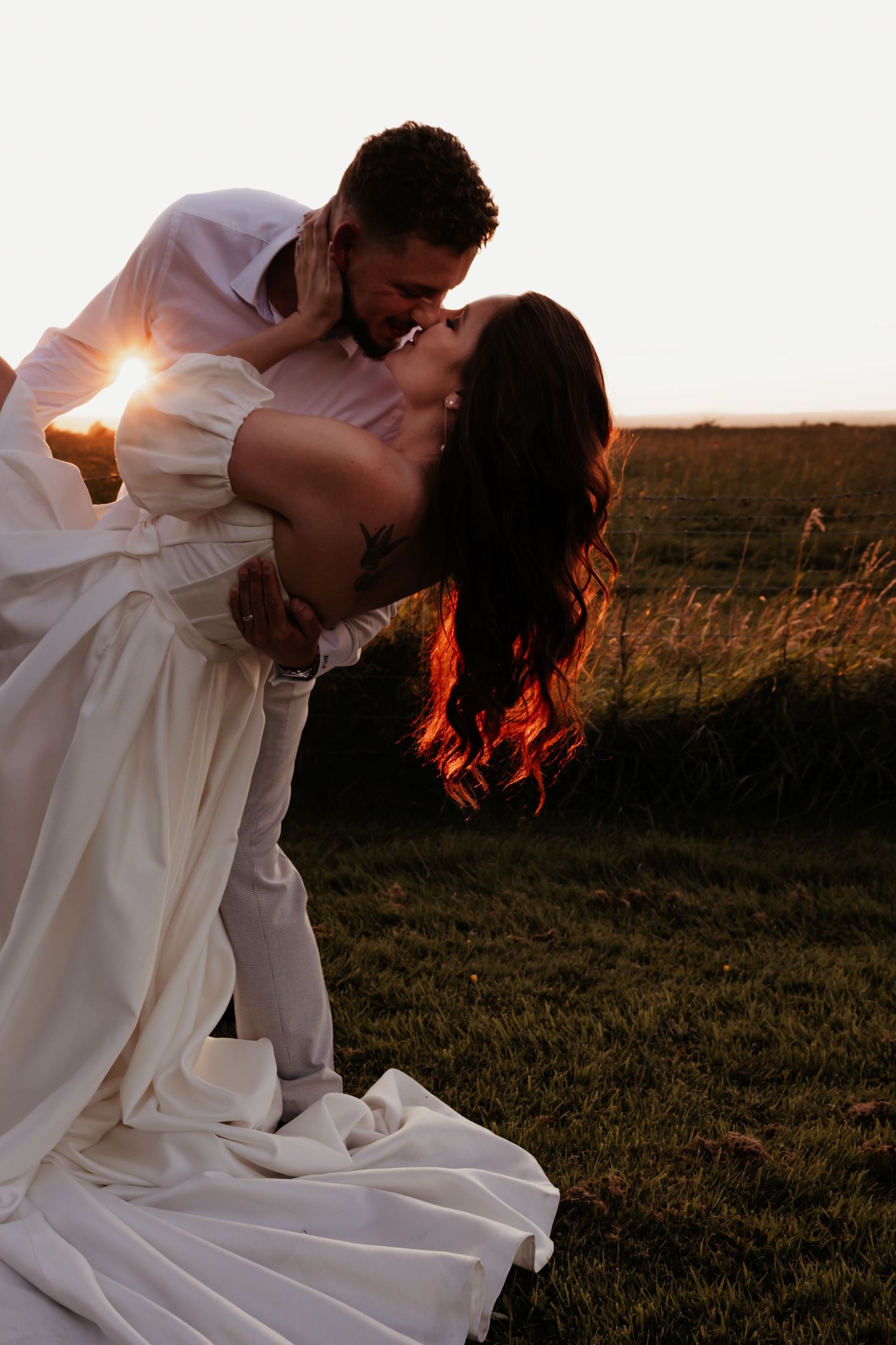 Romantic sunset dip kiss between couple in wedding attire with golden light behind them.