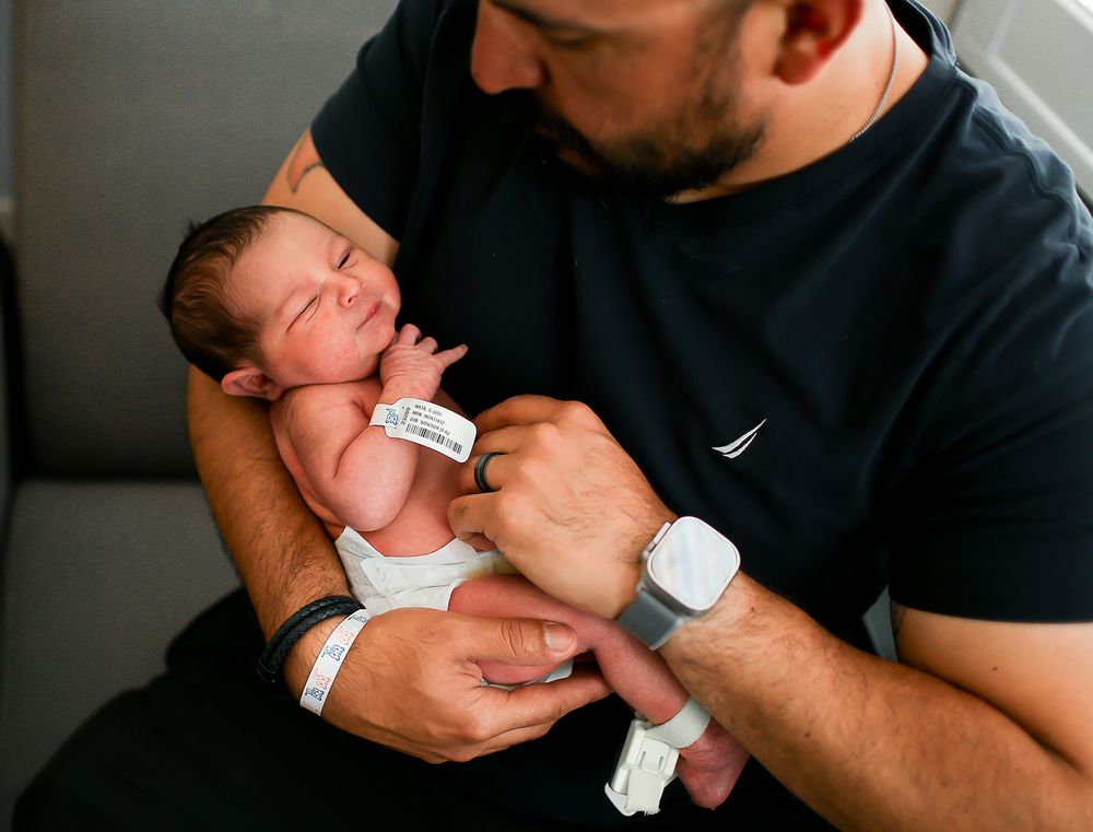 A newborn baby wearing hospital identification bands rests peacefully in caring arms on a gray couch.