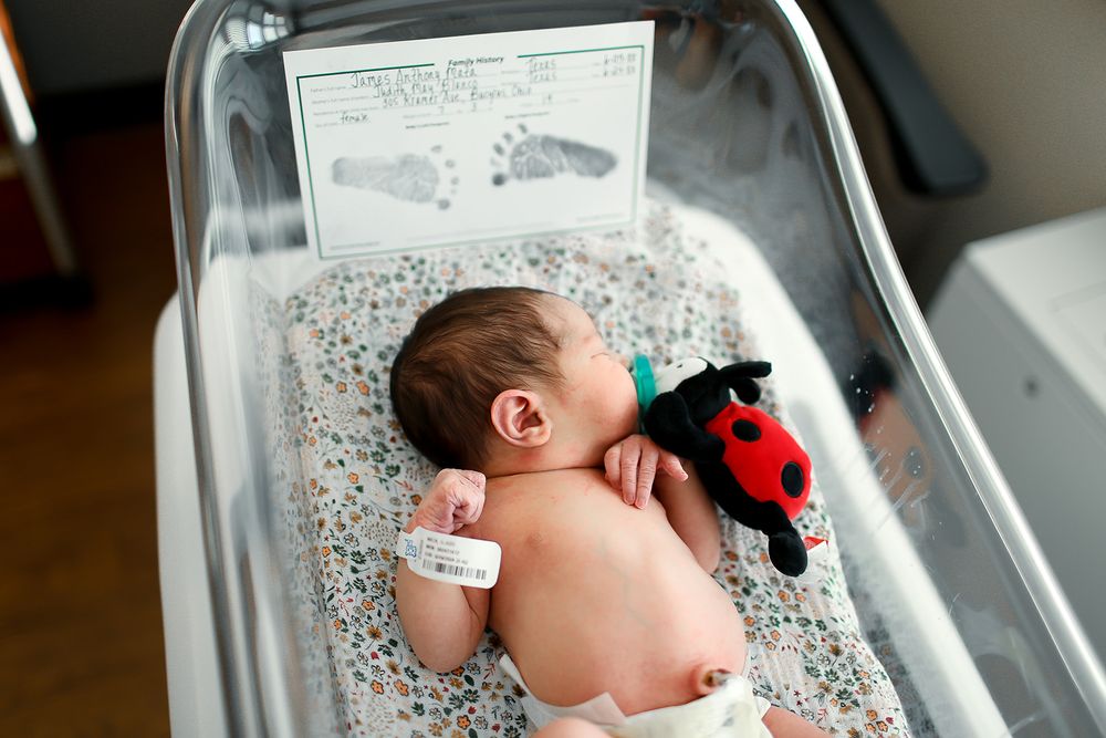 A newborn sleeps in a hospital bassinet with a stuffed ladybug toy and monitoring device visible.
