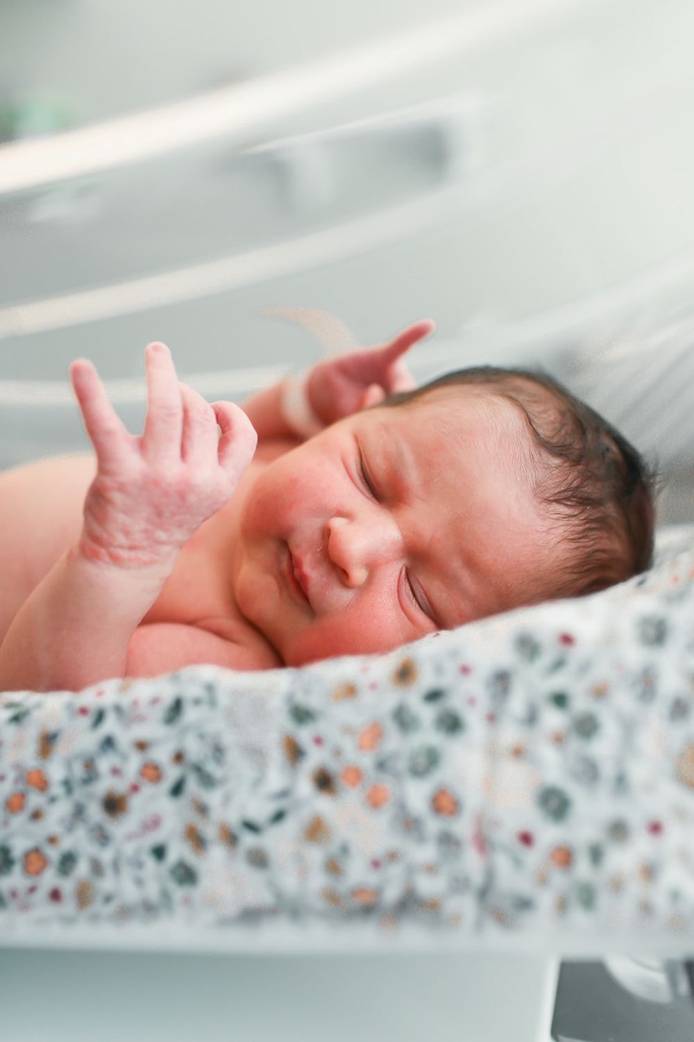 A newborn baby rests on a floral patterned blanket with hands raised and eyes gently closed.