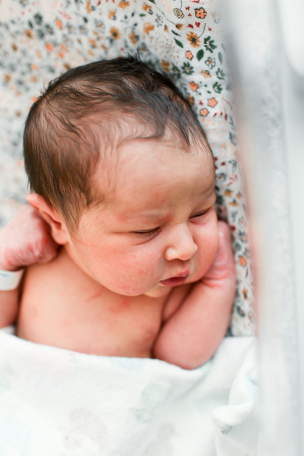 Sleeping newborn baby swaddled in delicate floral printed fabric rests peacefully against white bedding.