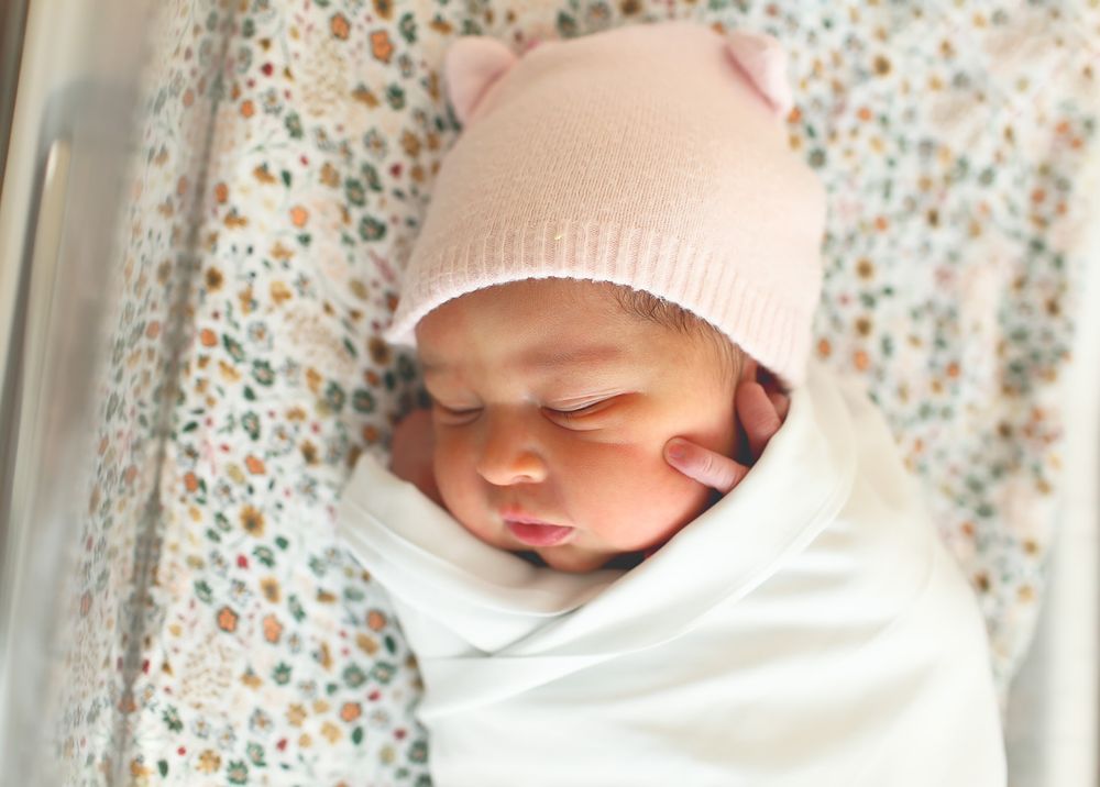 Sleeping newborn wrapped in white swaddle wearing soft pink knit beanie hat against floral pattern background.