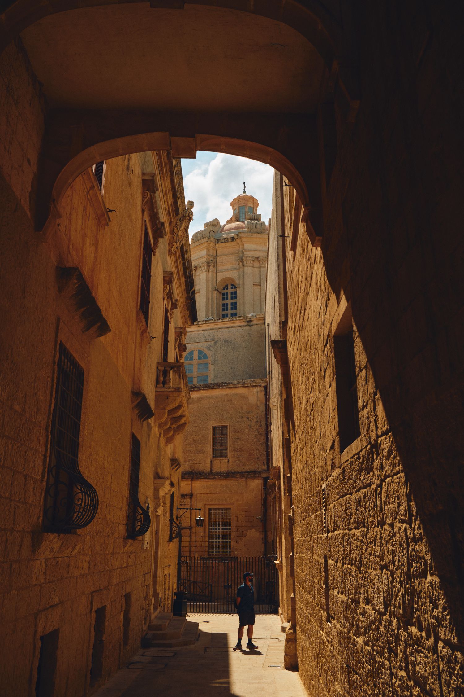 Narrow alley with stone walls leading to church dome through arched passageway.