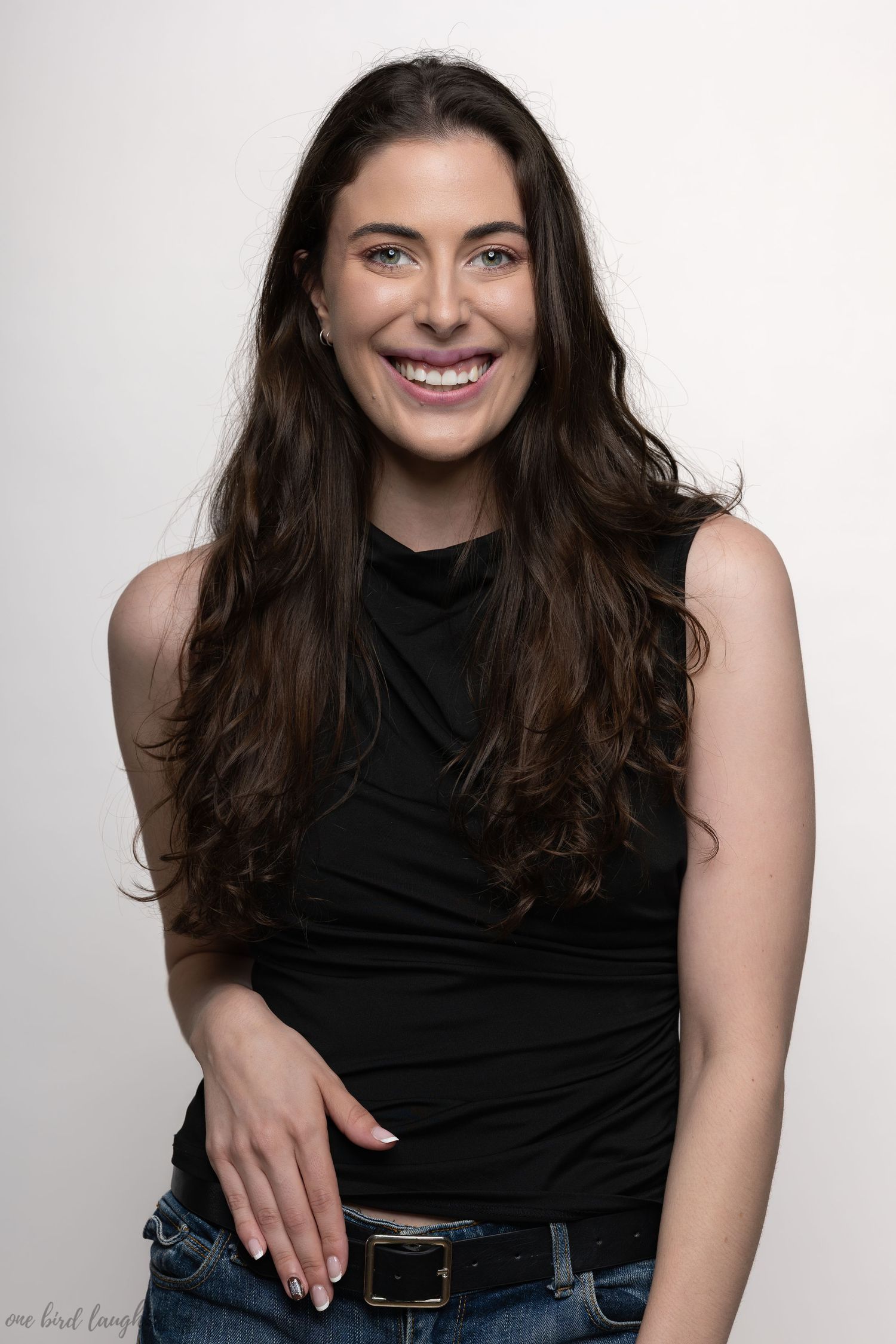 A person with long dark hair wearing a black sleeveless top and jeans poses against a light background with a warm smile.