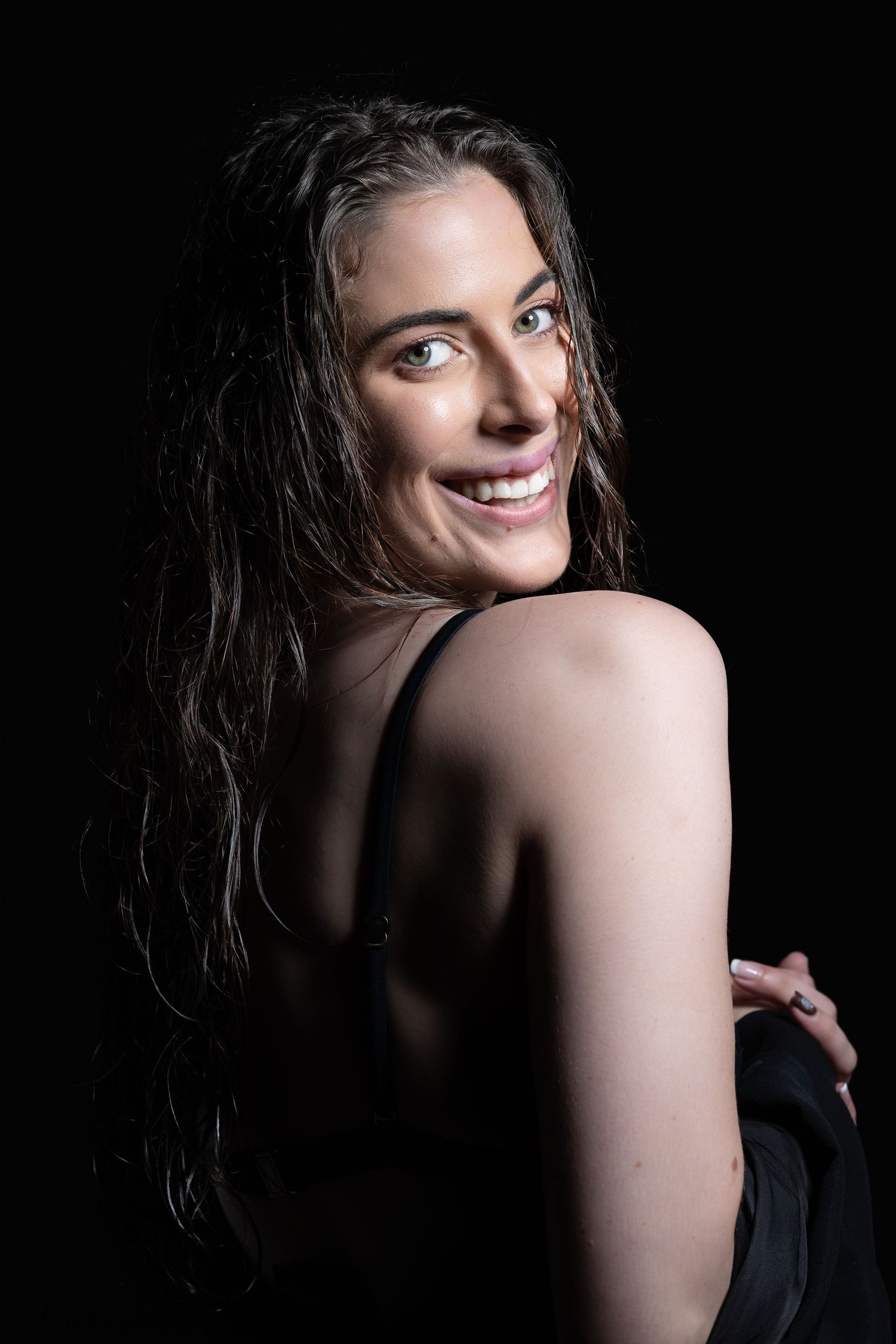 Dramatic portrait of a woman with long dark wet hair smiling over her shoulder against a black background.