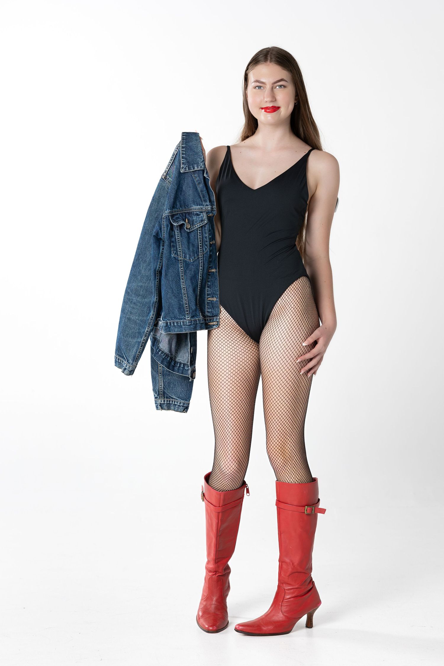 Fashion model wearing black bodysuit, fishnet stockings, red boots and denim jacket against white background.