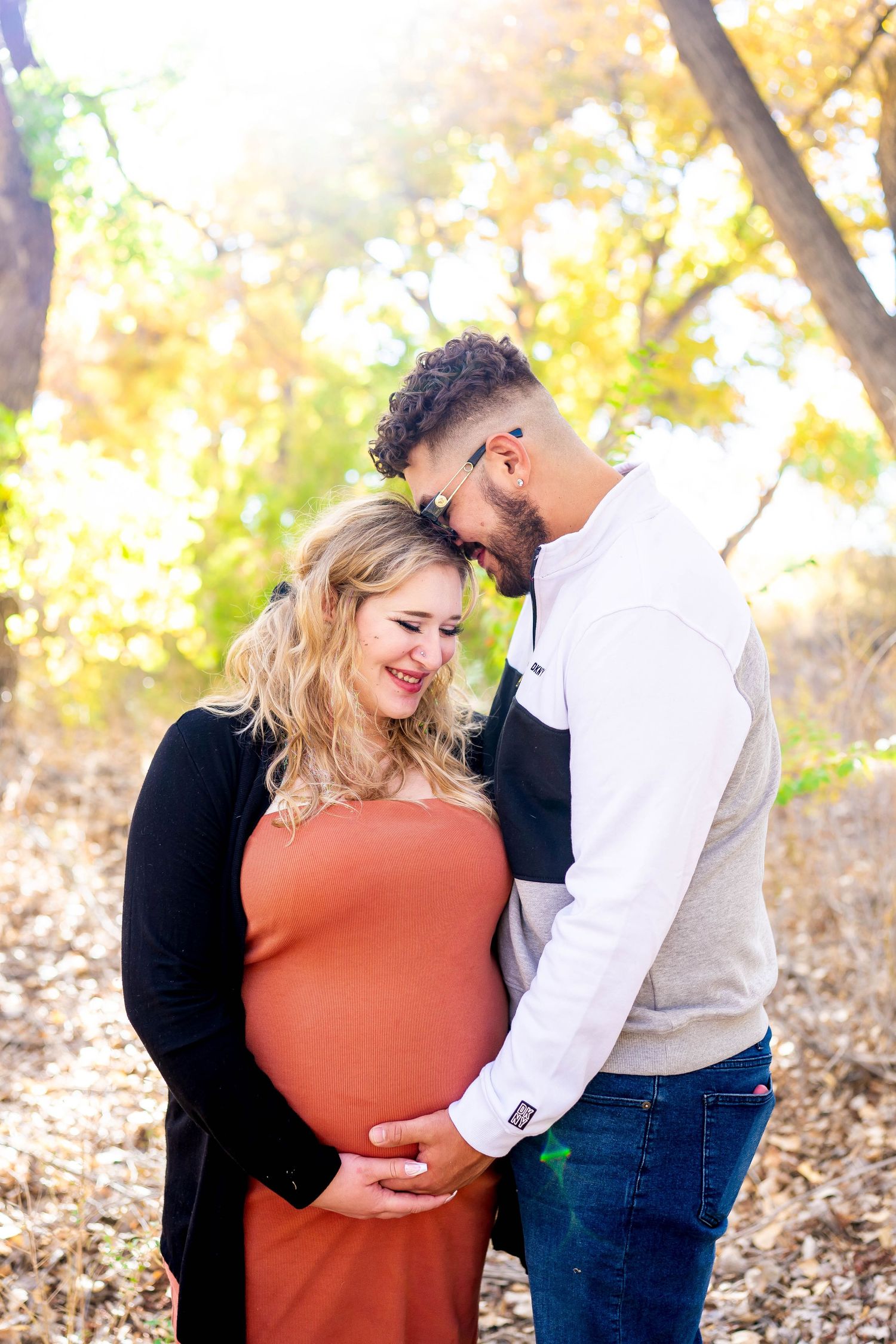 A tender maternity photo shows a couple embracing outdoors with autumn foliage in the background.