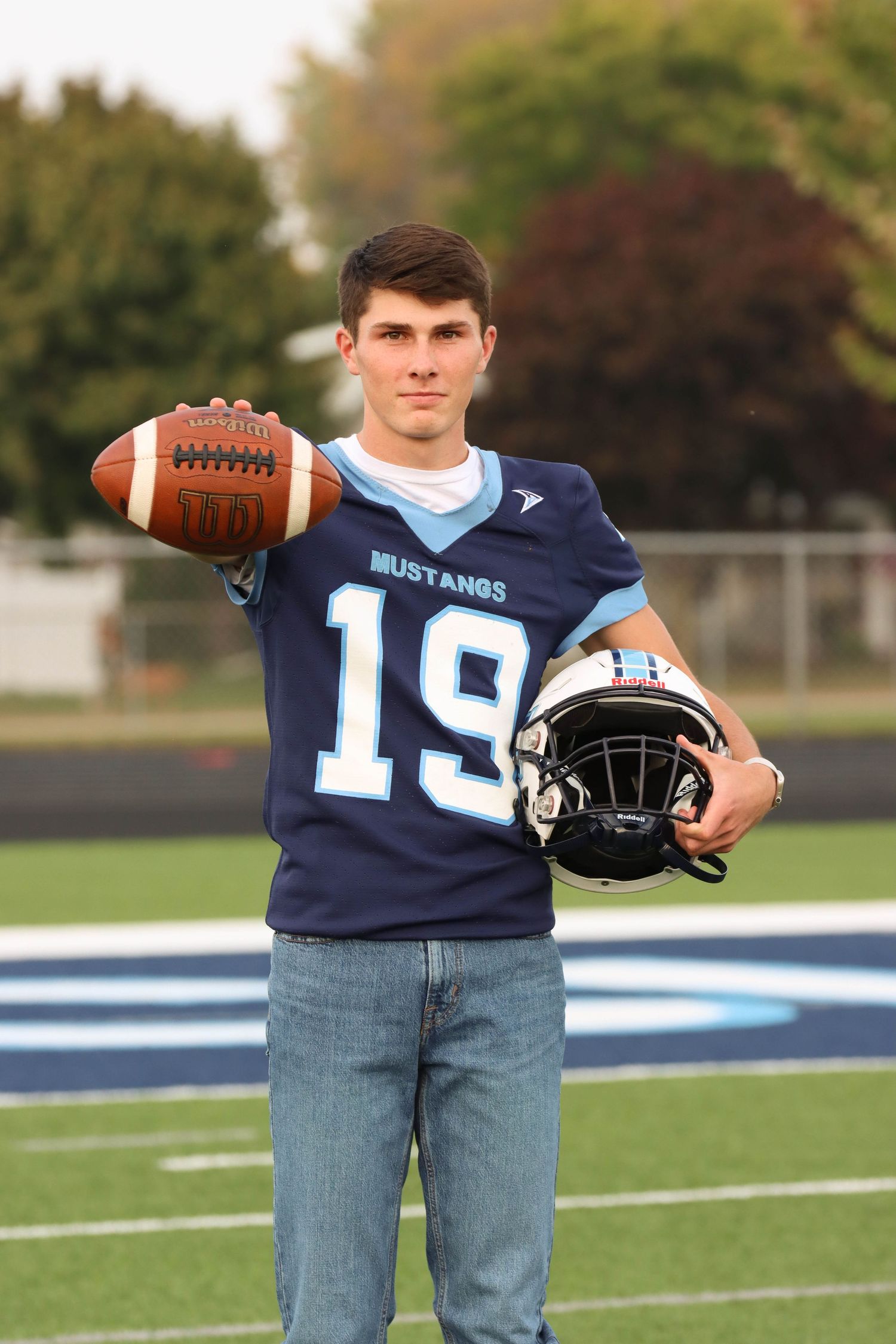 Football player holds ball and helmet on field wearing navy blue jersey number 19.