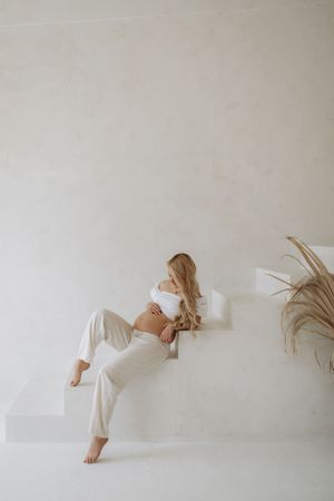 Minimalist maternity photo of woman in white loungewear reclining on stairs.