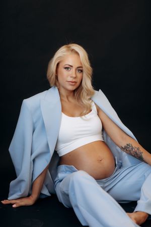 Maternity photos featuring woman in light blue blazer and white crop top against dark background.
