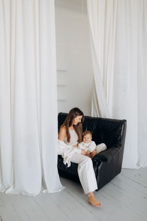 Mother and baby share tender moments on white couch in minimalist studio setting.