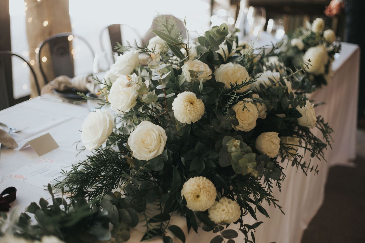 Elegant white roses and eucalyptus floral centerpiece arrangement on white linen wedding reception table.