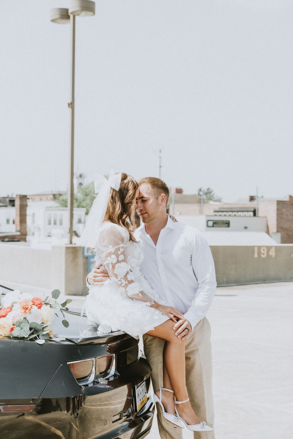 A couple in white attire share a tender moment against a black car on a sunny day.